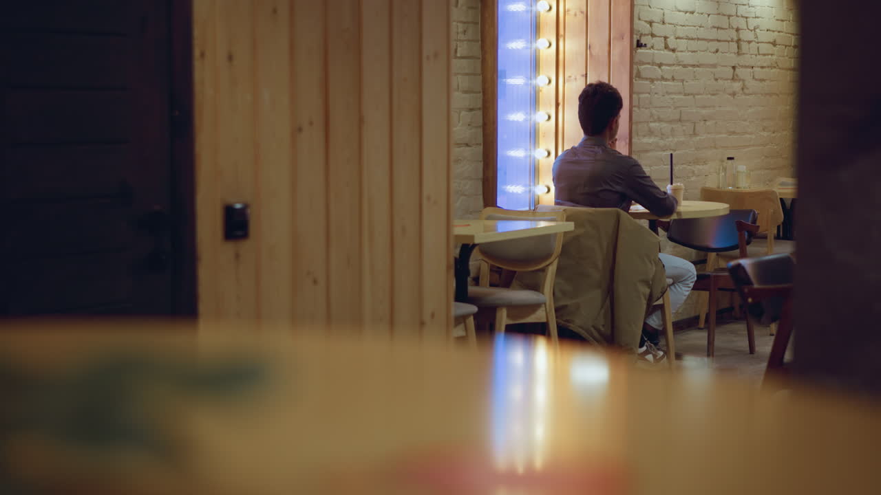 Man in gray shirt sitting alone near wooden wall and mirror lights in cozy empty caf , sipping from paper cup with straw, ambient lighting reflecting on tables, creating moody peaceful evening atmosphere