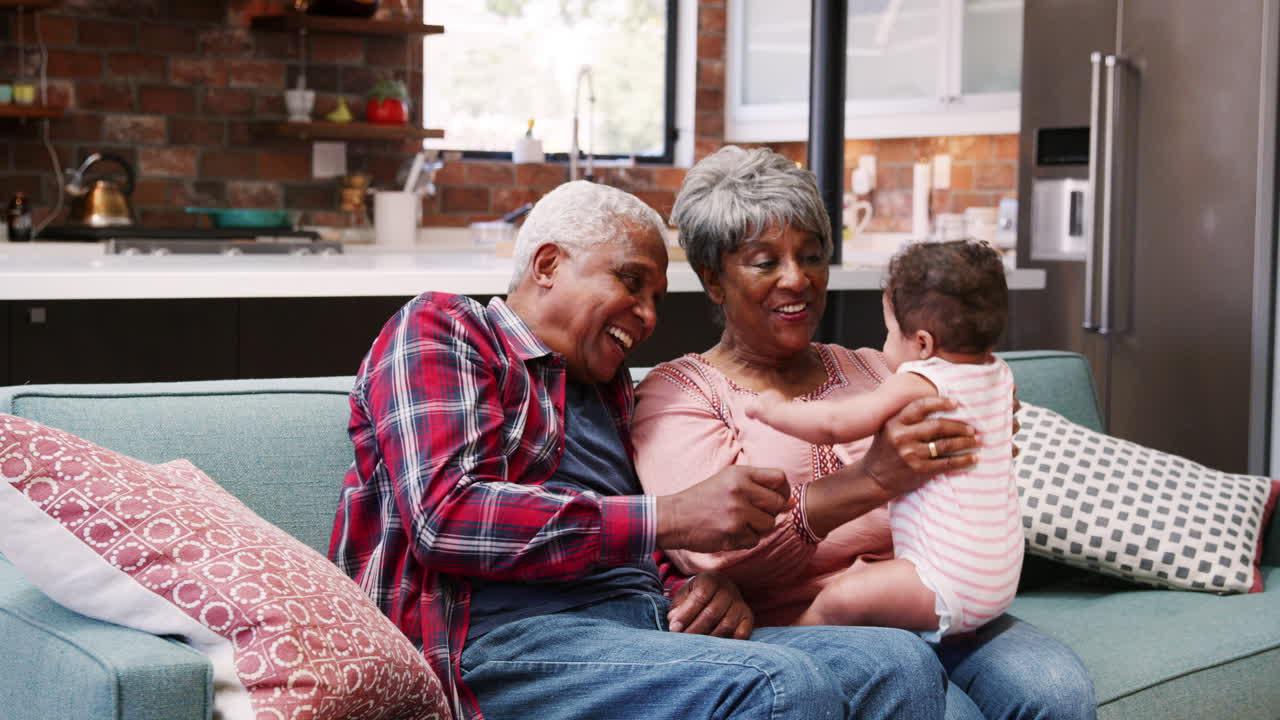 Grandparents Sitting On Sofa Playing With Baby Granddaughter At Home