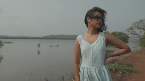 Slow-motion-panning-shot-of-an-attractive-young-woman-dressed-in-a-baby-blue-dress-standing-in-front-of-a-lake-during-windy-weather