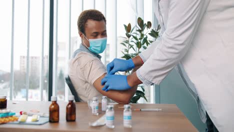 Primer-Plano-De-Un-Joven-Médico-Profesional-Con-Bata-Médica-Y-Protección-Que-Inyecta-La-Vacuna-A-Un-Paciente-Con-Máscara-Médica-Sentado-En-El-Centro-Médico-Para-La-Vacunación