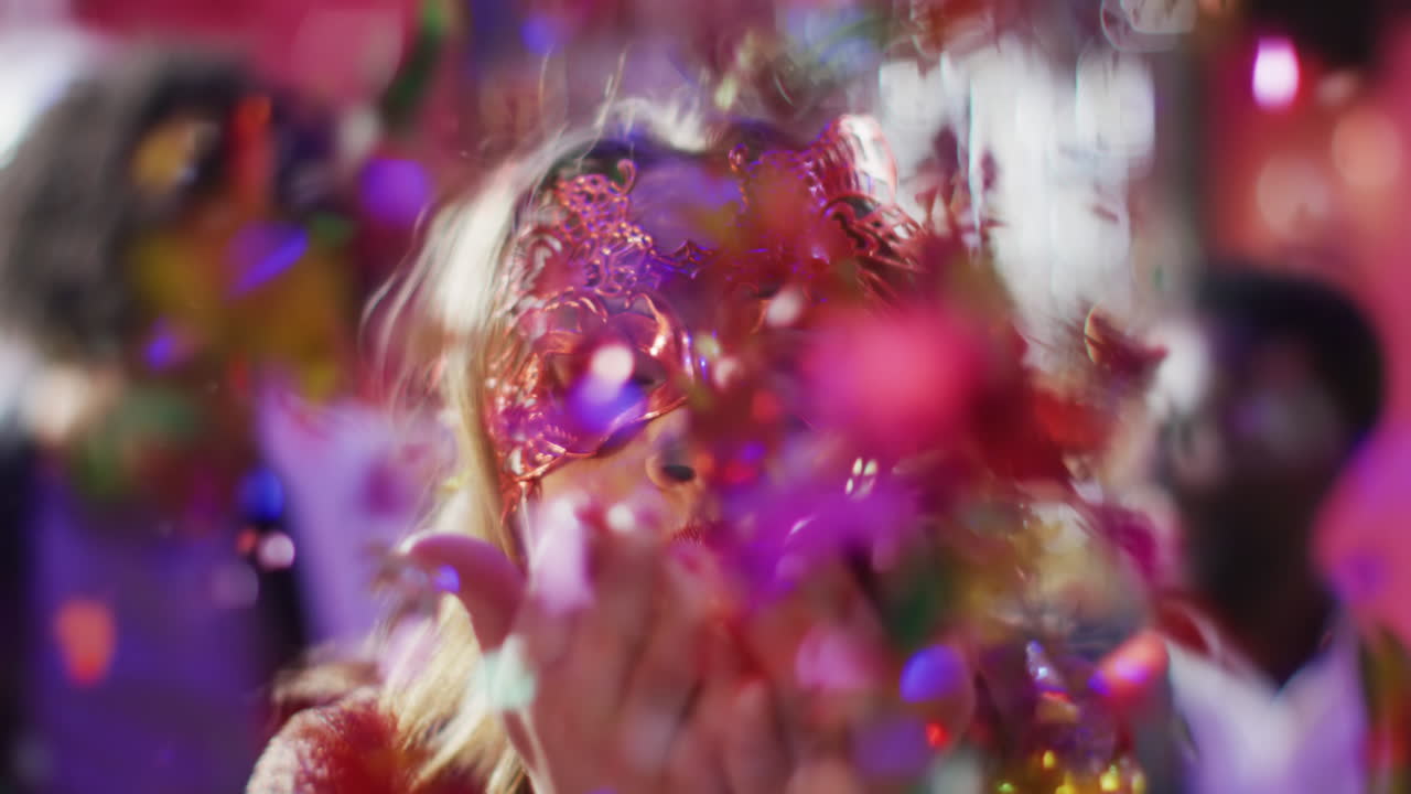 Video of happy caucasian woman in masquerade mask blowing glitter to camera at a nightclub