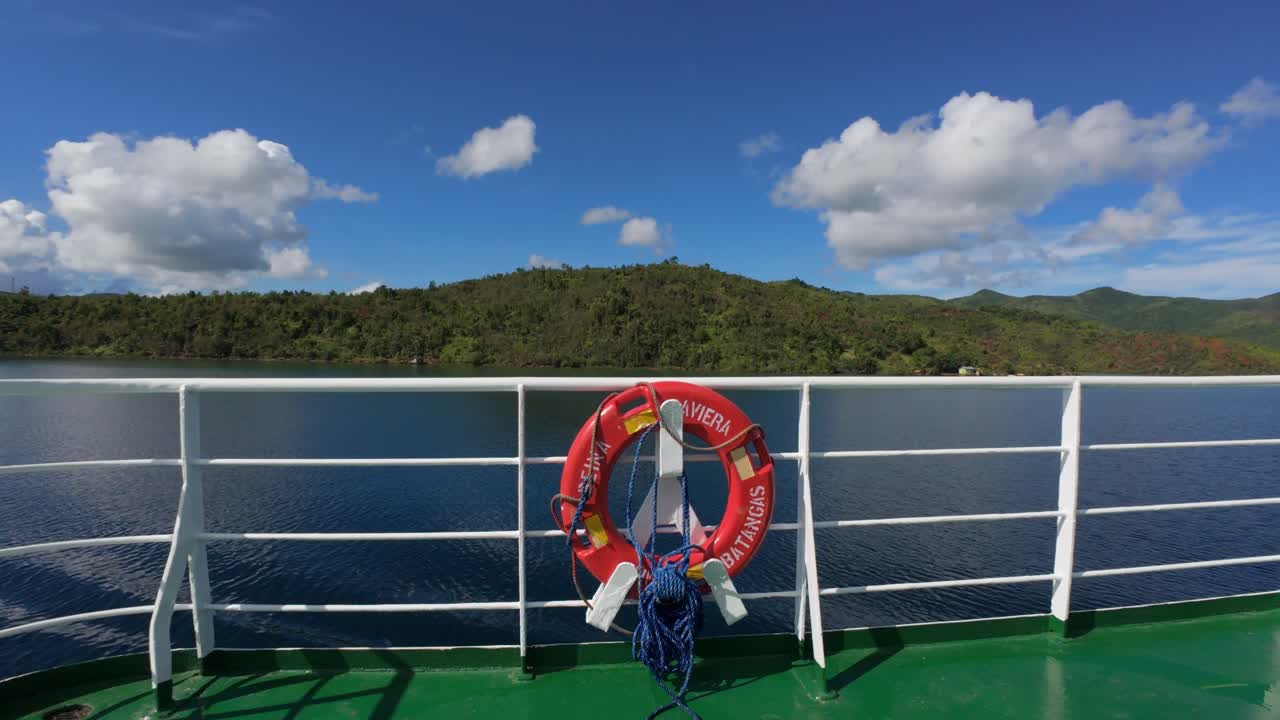 Lifebuoy hanging on ferry railing with calm sea and hills in background.