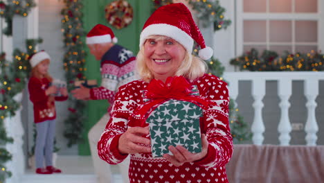 Portrait-of-grandmother-woman-presenting-gift-box-smiling-near-decorated-Christmas-house-with-family