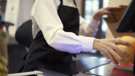 unrecognizable slender saleswoman in white shirt and black apron scanning product, fruits at checkout counter in store and putting it into brown paper bag. close up