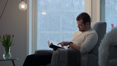 Man-sits-by-window-reading-book,-snowing-outside