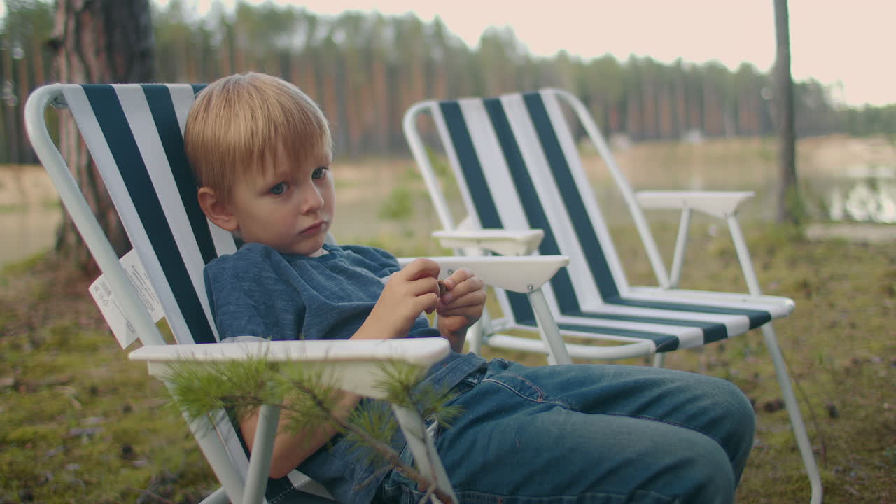 un niño aburrido está sentado en una silla en la naturaleza viajando y acampando en las vacaciones de verano descansando y relajándose