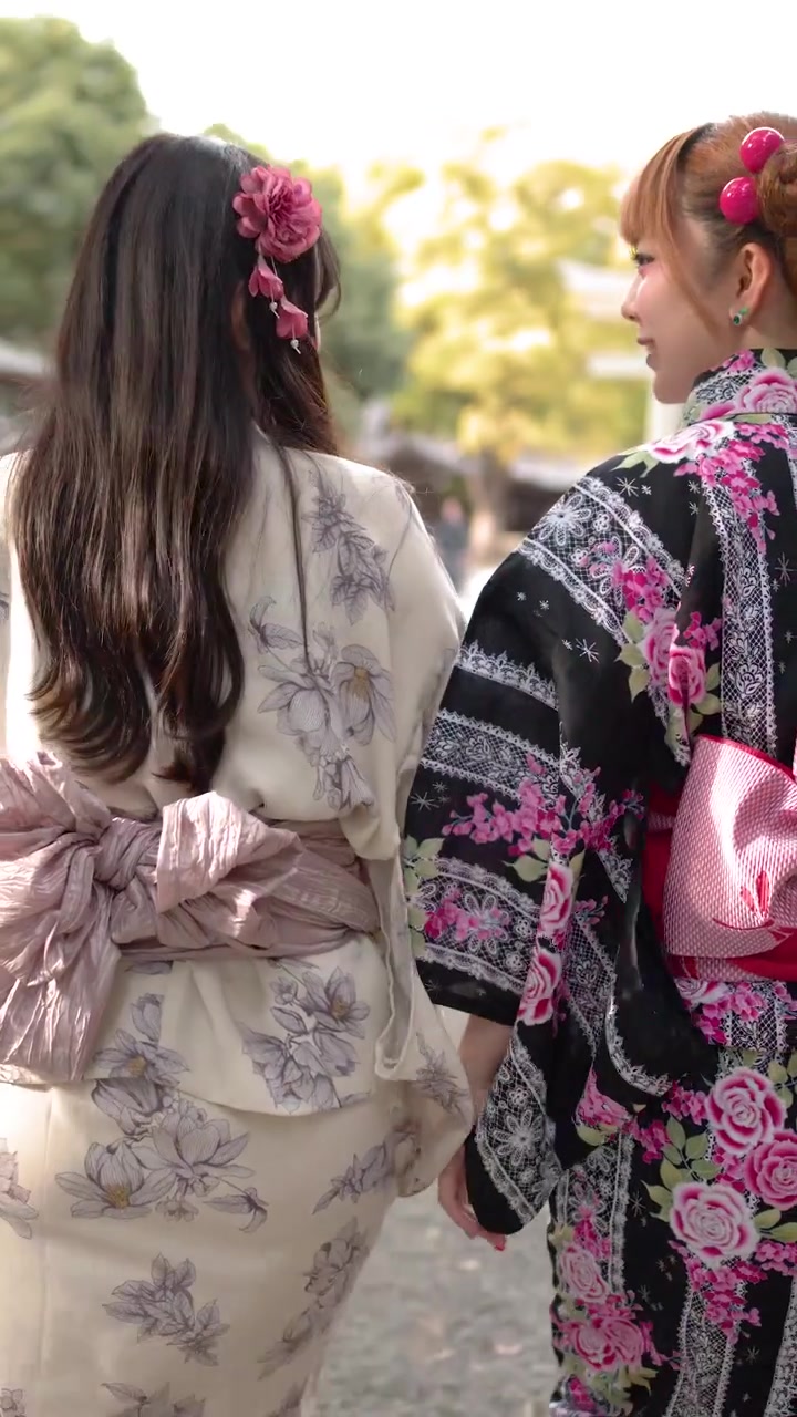 花柄の着物を着た2人の女性が、陽光が降り注ぐ日本の神社の庭園を歩く後ろ姿