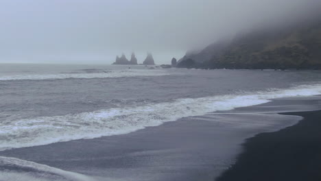 panorámica cinematográfica aérea hacia adelante desde las olas de la playa que se estrellan en vik islandia niebla temprana de invierno en la playa de arena negra
