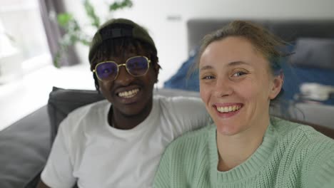 pareja interracial alegre tomando selfies con el teléfono inteligente mientras están sentados juntos en el sofá