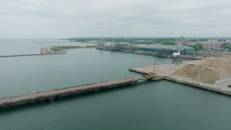 aerial establishing view of port cranes and empty loading docks at port of liepaja , liepaja city in the background, overcast summer day, wide drone shot moving forward