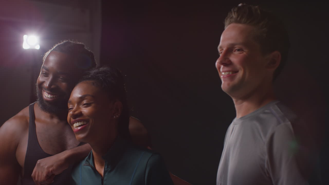 Studio Portrait Of Smiling Male And Female Athletes In Fitness Clothing Having Fun Posing And Flexing Muscles Against In Gym With Low Key Lighting