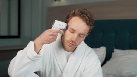 Un-Joven-En-Bata-De-Baño-Medirá-Su-Temperatura-Con-Un-Termómetro-Sin-Contacto