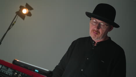 focused keyboardist in black hat and outfit plays a red sampler in a moody grey studio