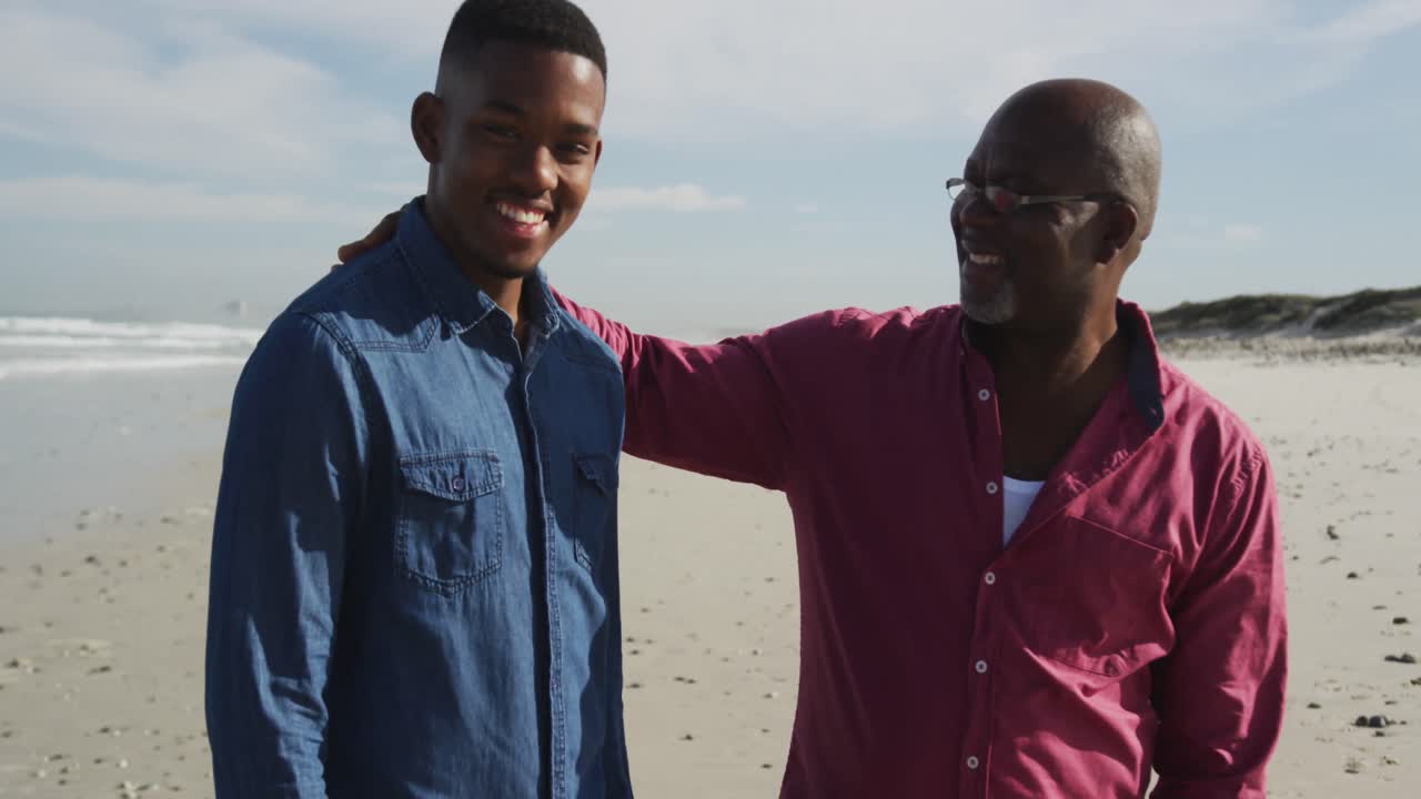 African american father standing on beach with teenage son, putting hand on his shoulder and smiling