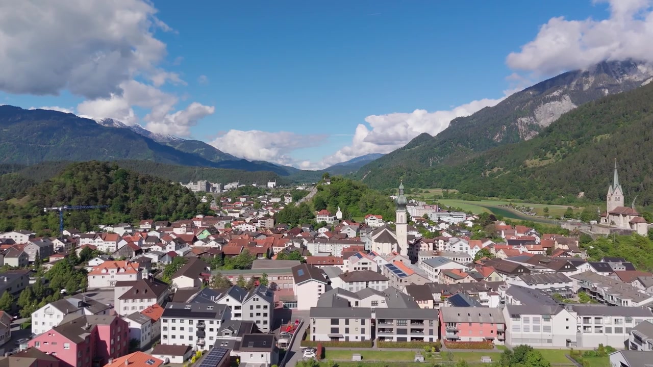 Vista aerea con drone di Domat Ems, Svizzera, con il centro città, i campanili delle chiese e le montagne alpine circostanti sotto un cielo azzurro brillante