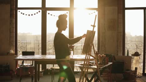 Fully-concentrated-sophisticated-female-artist-in-her-20's-is-drawing-picture-on-easel.-The-sun-behind-lits-up-the-art-studio-and-turns-it-into-inspiring-environment-of-creativity.