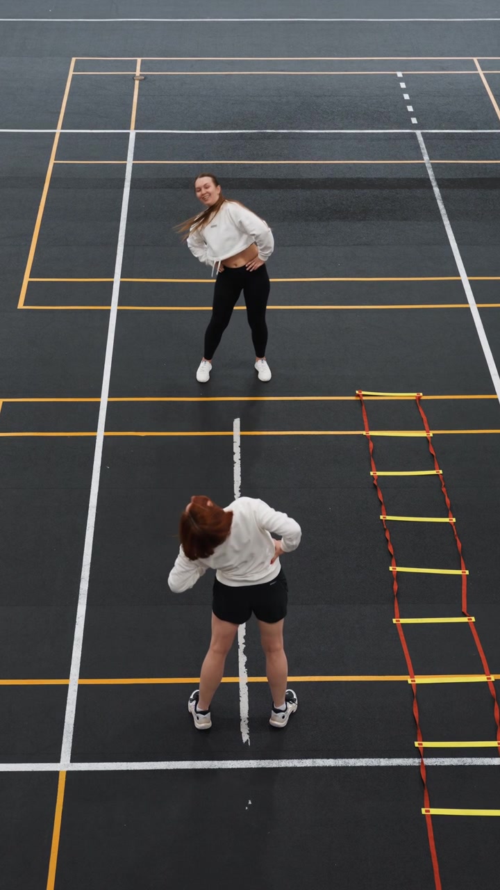 Coach and trainee practice badminton on court, topdown composition showing instructor guiding concentrated pupil through ladder agility and footwork drills, handsonhips stance, bent posture, marked