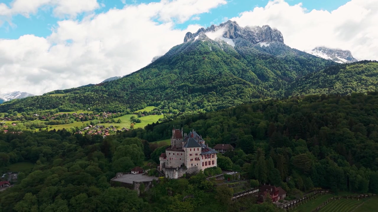 Tomada aérea ao redor do castelo de Menthon Saint Bernard com os Dents de Lanfon ao fundo em um dia ensolarado de primavera, departamento de Haute Savoie, região de Auvergne-Ródano-Alpes, França.