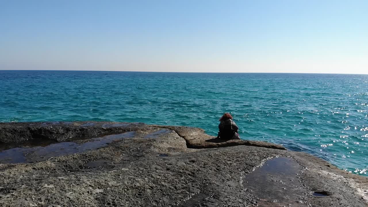 바람이 머리를 불고 푸른 물을 바라보며 바위에 앉아 있는 소녀의 공중 추적 샷