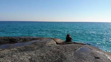 Luftaufnahme-Eines-Mädchens,-Das-Auf-Einem-Felsen-Sitzt-Und-Auf-Blaues-Wasser-Blickt,-Während-Der-Wind-Ihr-Die-Haare-Weht