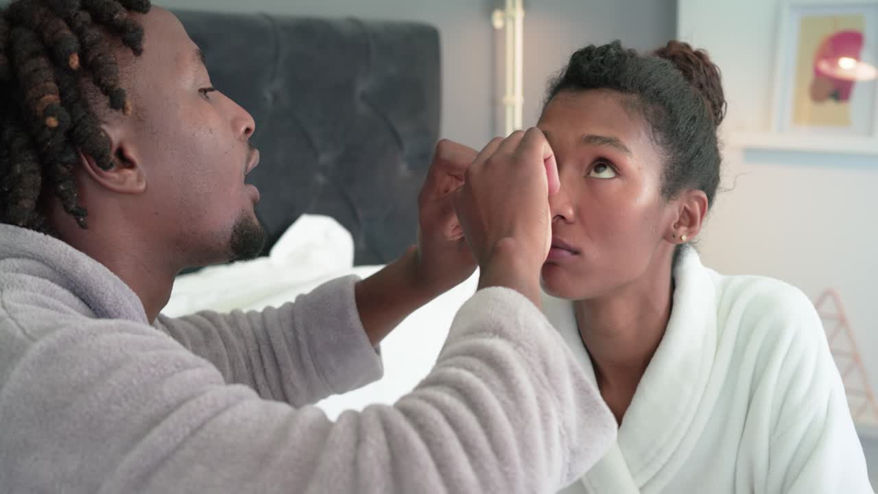 African American couple sitting on bed, man offering blue dropper, using white tool, placing lens