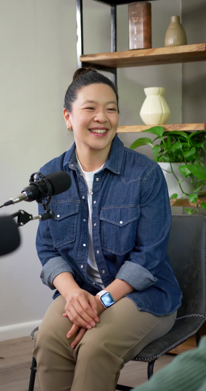 Vertikal video: Vært hilser, asiatisk kvinde i denimskjorte smiler, nikker nær mikrofon til podcast.