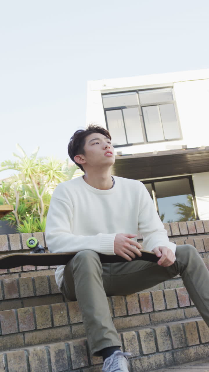 정원에서 스케이트보드와 함께 앉아있는 아시아 남성 십대의 수직 비디오