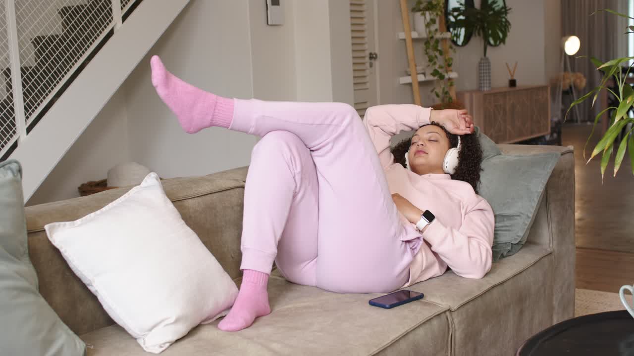 African American woman reclining on couch, lifting right leg, adjusting headphones for fit, smiling