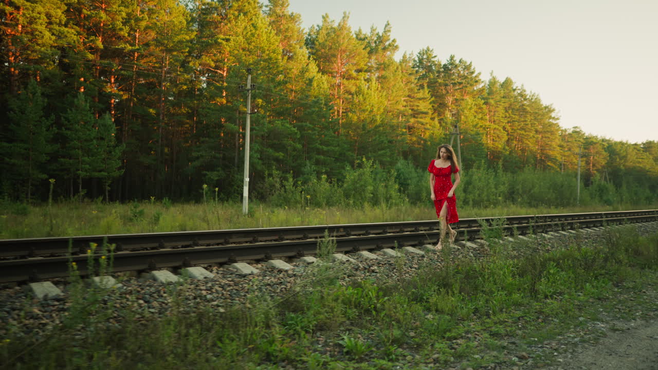 Young lady in red dress jumping carefully between railway sleepers while holding gown for balance, surrounded by forest trees, utility poles, gravel and wild grass under warm golden sunlight