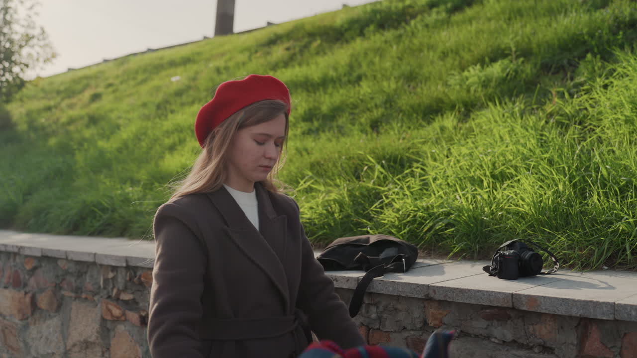 Kaukasisk kvinde folder rødt ternet tørklæde på havnepromenade nær græsklædt skråning, iført rød baret og brun frakke, hænder glatter uldstof over stenmur, vintagekamera og taske i nærheden, nærbillede