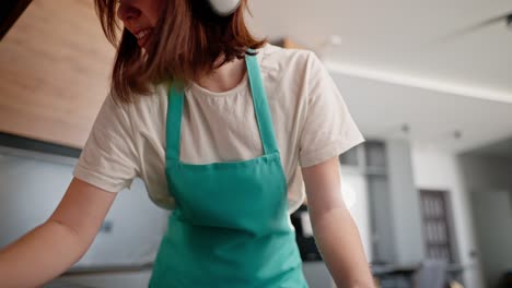 Eine-Brünette-Putzfrau-In-Einem-Weißen-T-Shirt-Und-Einer-Blauen-Schürze-Wäscht-Den-Herd-In-Der-Küche-Eines-Modernen-Studio-Apartments-Und-Hört-Zu-Hause-Mit-Kabellosen-Reinigungskopfhörern-Musik