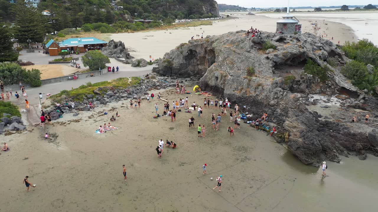 Circling drone shot of people dancing on the beach next to large rock formation.