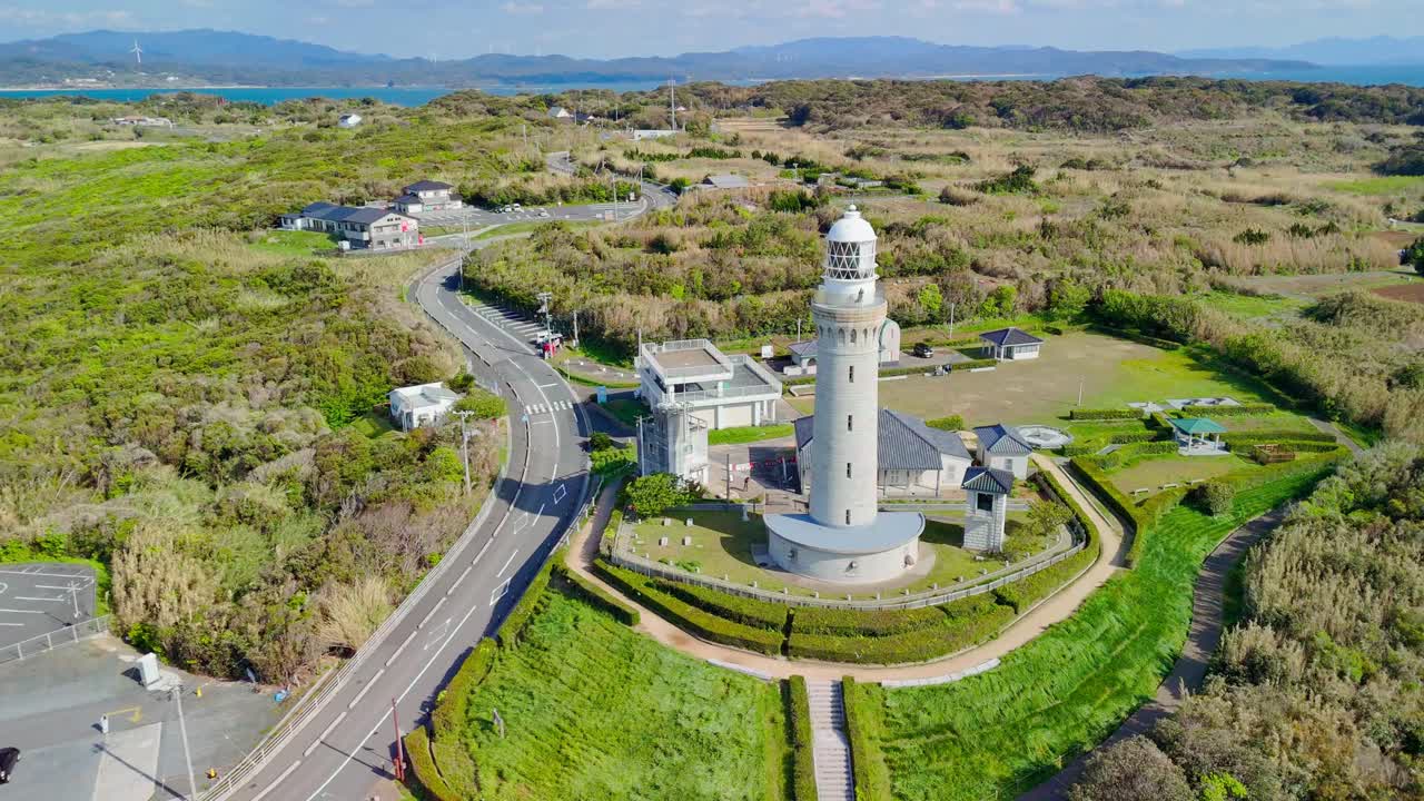 A stunning aerial drone shot pulling back from the historic Tsunoshima Lighthouse to reveal the entire complex, nearby road, and lush green coastal landscape of Yamaguchi, Japan