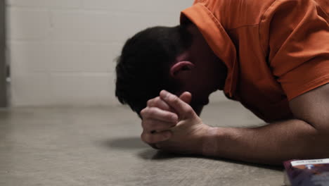 a prison inmate lays on ground crying, praying, weeping, sobbing next to bible while wearing orange prison uniform jumpsuit