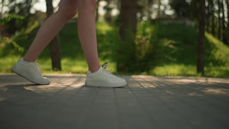 side leg view of individual wearing white sneakers walking confidently on interlocked pathway next to grassy field under warm sunlight, with blurred background featuring trees casting soft shadows