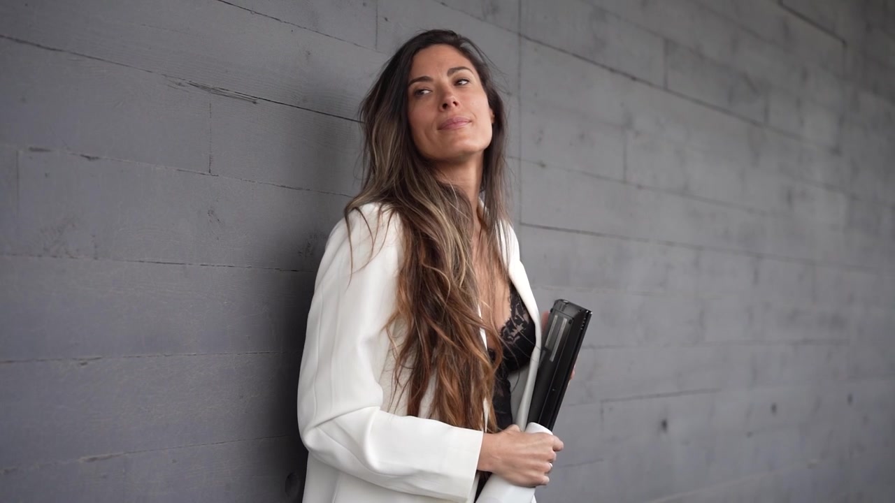 Uma mulher com cabelos longos e esvoaçantes está segurando um caderno enquanto está encostada em uma parede cinza.