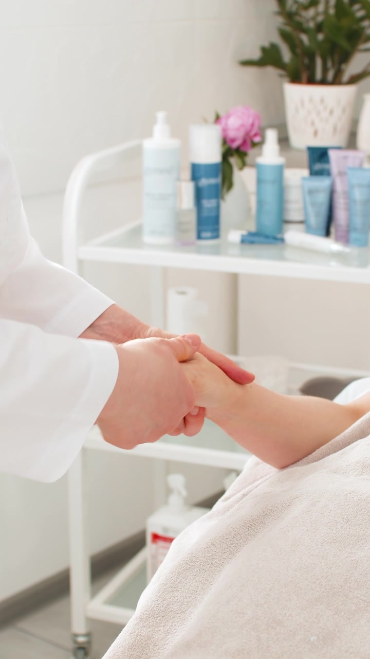 Kosmetolog giver en kvinde afslappende håndmassage under en ansigtsmaskebehandling i en skønhedsklinik og spa.