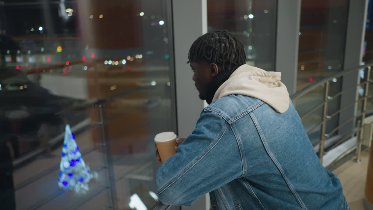 Young man in denim jacket and hoodie holding coffee cup while leaning on railing, gazing through large window at night city lights, inside modern building with glass panels and warm interior lighting