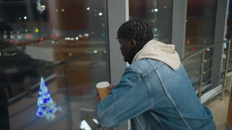 young man in denim jacket and hoodie holding coffee cup while leaning on railing, gazing through large window at night city lights, inside modern building with glass panels and warm interior lighting