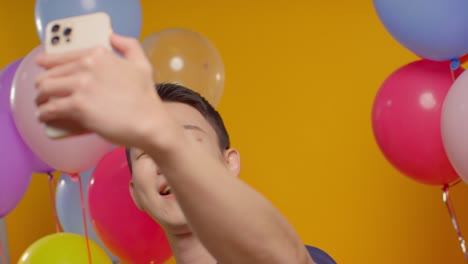 Studio-Portrait-Of-Man-Taking-Selfie-Wearing-Party-Hat-Celebrating-Birthday-Surrounded-By-Balloons-2