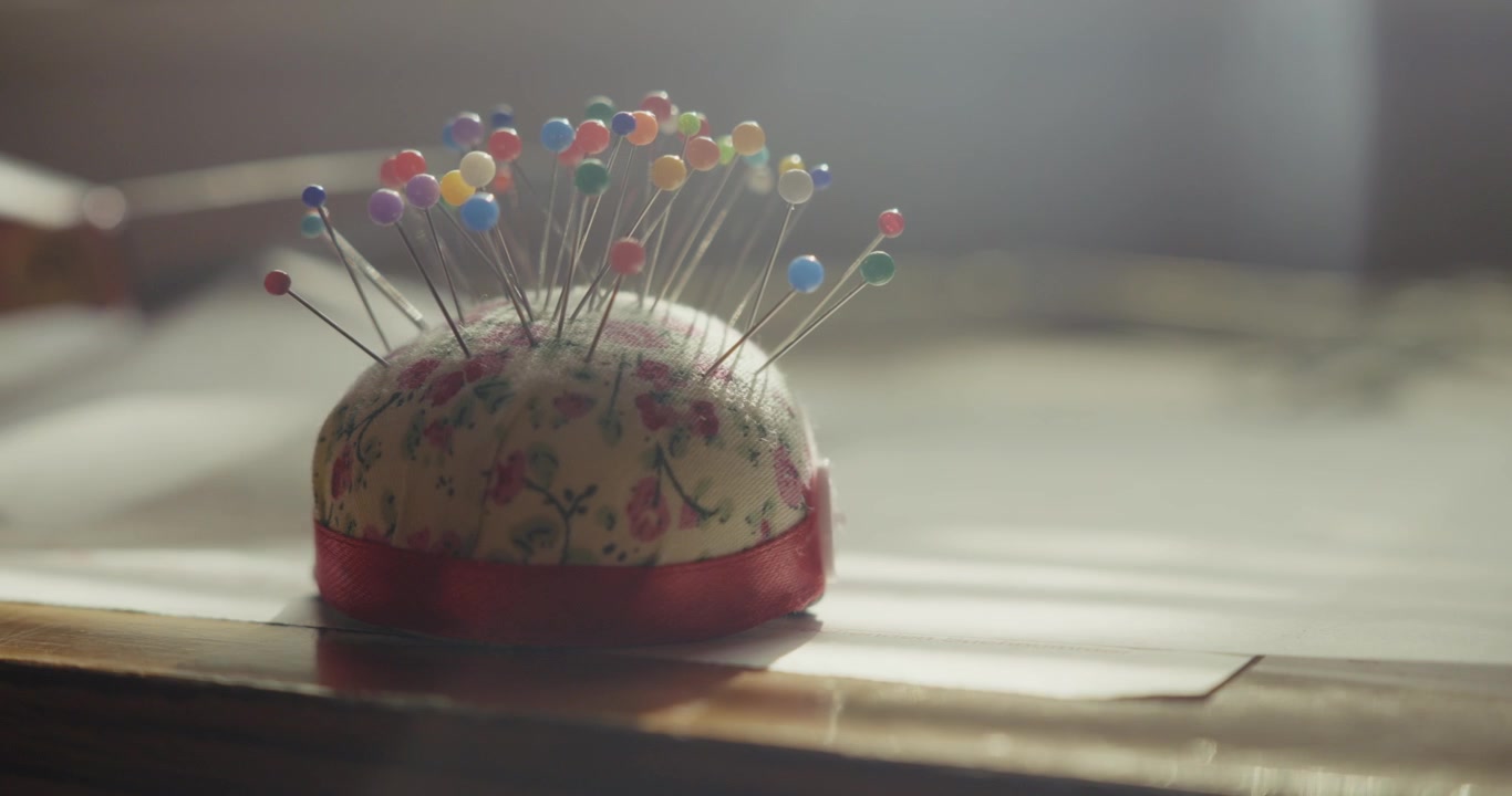 Close-up of a hat pin cushion in soft light while a fashion designer works cutting fabric in the background of a tailoring studio.