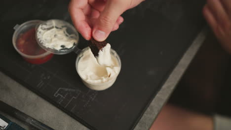 close up of fair skinned man dipping dark snack into creamy white sauce holding it carefully with fingers next to container of red sauce and creamy sauce on black mat in casual workspace environment