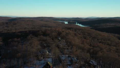 Drone-flyover-rural-mountain-homes-in-a-forest-towards-a-gorgeous-lake-in-the-distance,-at-winter-sunrise
