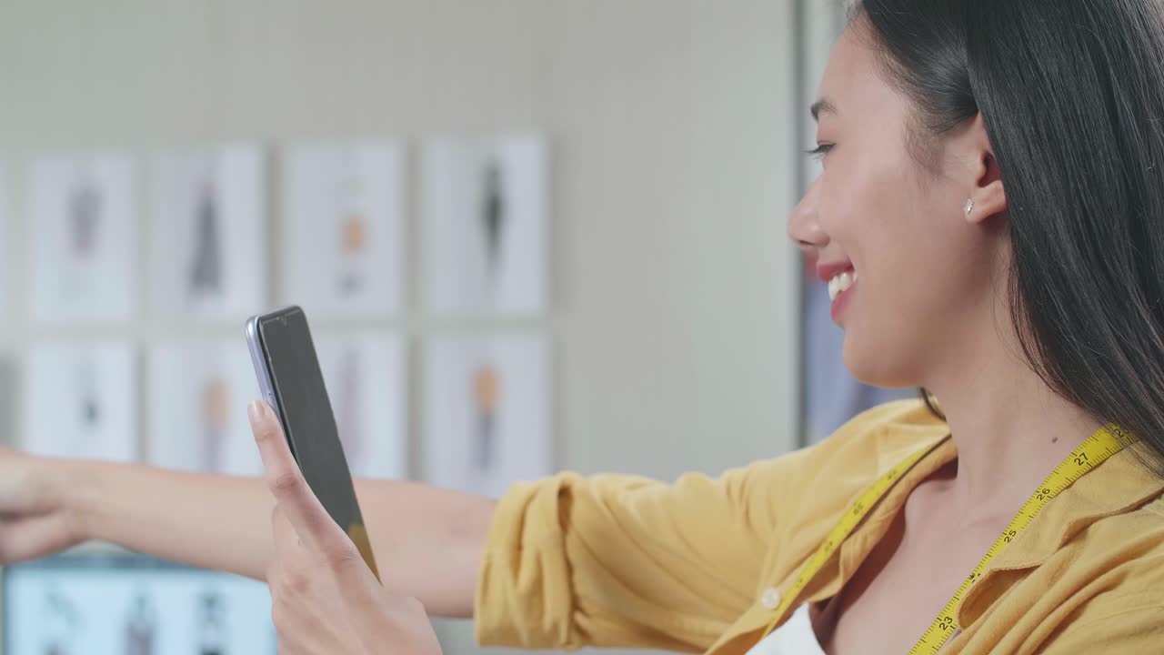 웃는 아시아 여성 디자이너가 스마트폰을 사용하여 스튜디오에서 옷을 디자인하는 동안 마네킨에 드레스의 사진을 찍습니다.