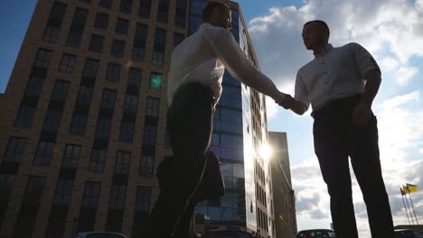 two successful businessmen meeting in city and greeting each other. male colleagues shaking hands in urban environment. handshake of business partners outdoor. cityscape at background. low view