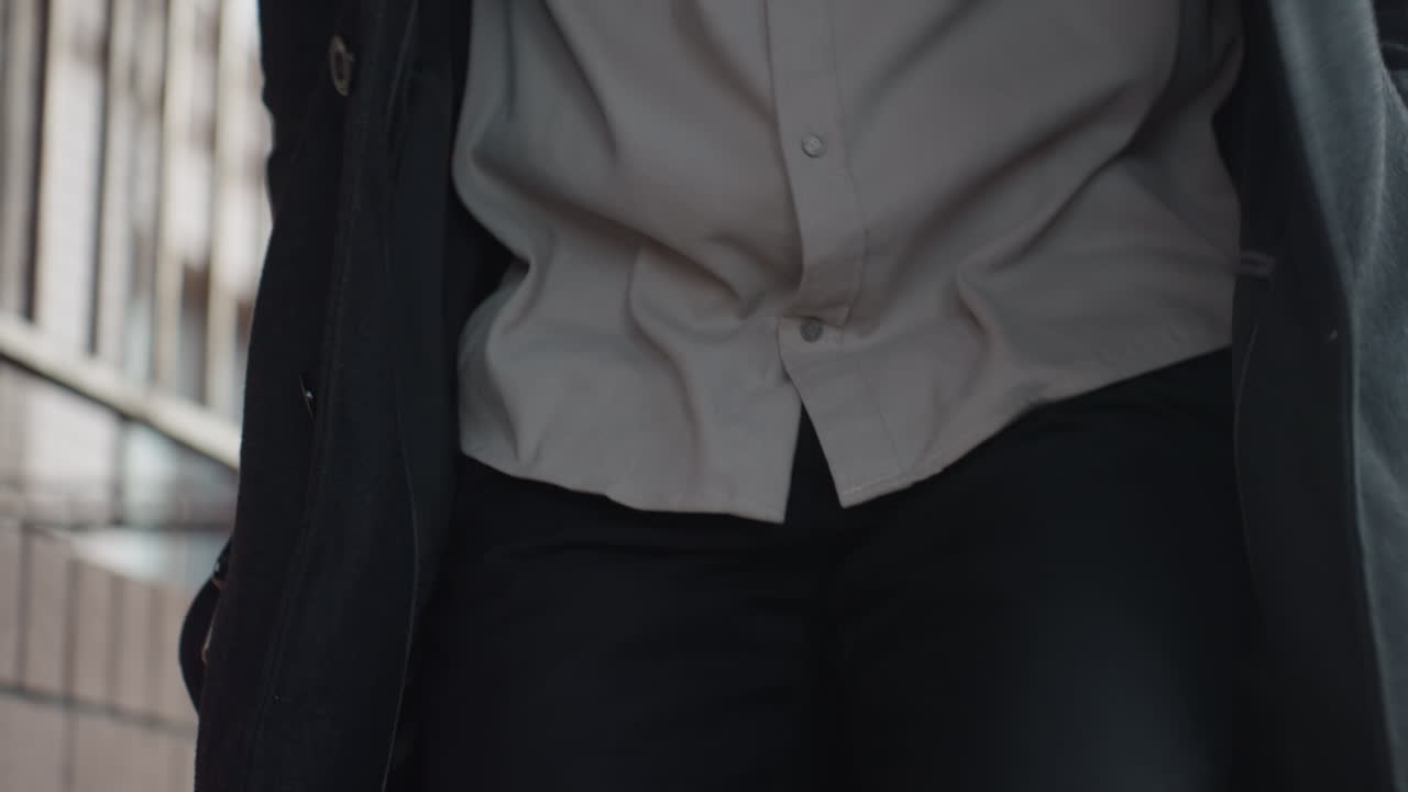 클로즈업 토르소 뷰는 베이지색 셔츠가 어두운 코트 아래에서 펄럭이는 모습을 보여주고, 손은 난간을 지나 걸어가면서 주머니 속으로 들어가고, 검은색 바지가 프레임 안에 있으며, 부드러운 초점 배경에는 벽돌과 덩굴 격자가 보여요.