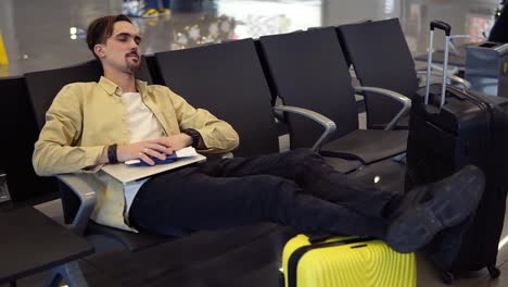 un hombre barbudo con camisa amarilla durmiendo con las piernas en la maleta amarilla, sosteniendo sus cosas en la sala del aeropuerto mientras