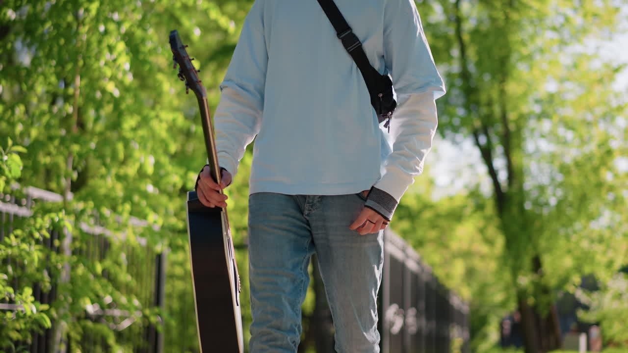 Giovane uomo che cammina con custodia di chitarra in un parco soleggiato, indossa un maglione bianco e jeans, tracolla sul petto, porta uno strumento acustico al suo fianco, passo rilassato oltre una recinzione di legno e verde