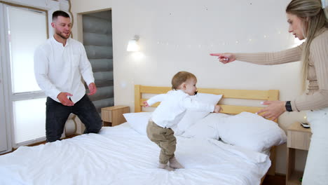 blonde woman holds her little baby above the head and puts on the bed. kid stands up and points at something. father stands at the bed.