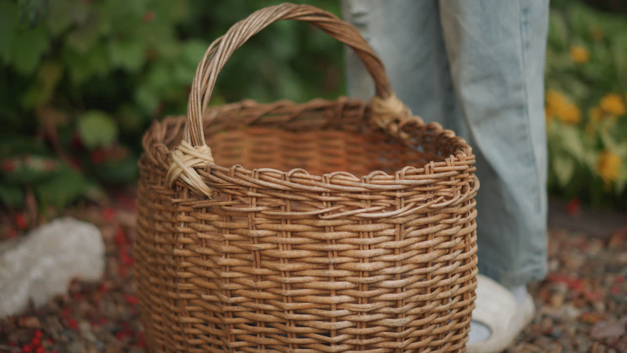 Cesta de vime sobre cascalho ao lado dos pés, jardineiro em pausa de jeans e chinelos, alça trançada repousando perto de um caminho de pedras, folhas espalhadas e luz natural suave, um clima rústico de colheita caseira.